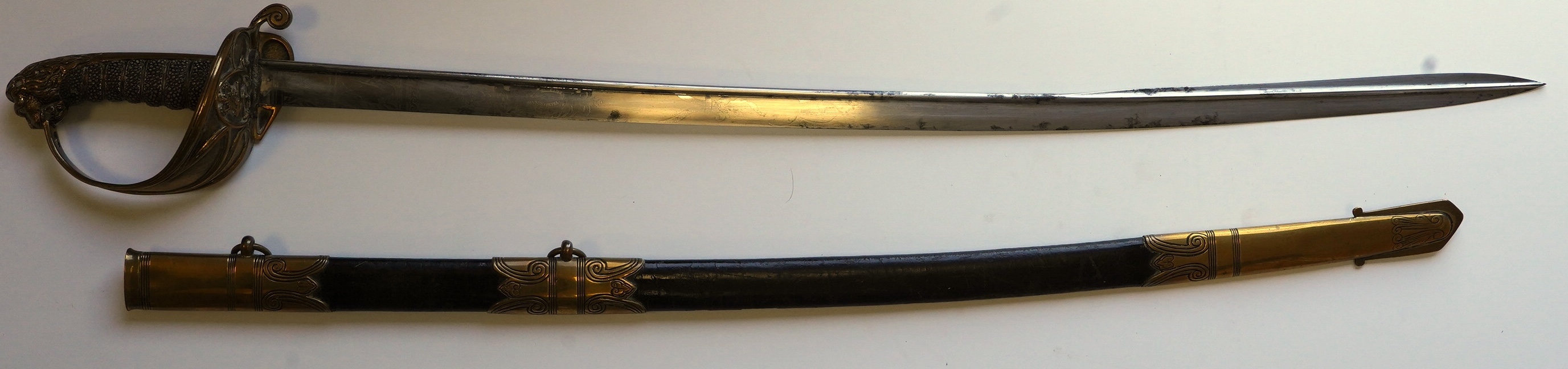 An early 19th century Royal Navy officer's sword with engraved blade, folding knuckle guard, lion’s head pommel and shagreen grip, leather scabbard with brass fittings, blade 73.5cm. Condition - fair, some wear overall.